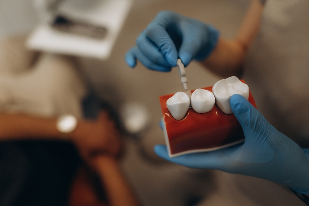 A person in blue gloves holds a model of teeth, demonstrating a dental procedure with a tool. Another individual is blurred in the background. The environment resembles a dental office.