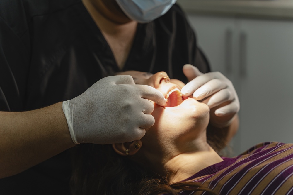 A dentist wearing gloves and a mask examines a patient’s mouth in a brightly lit dental office. The patient, in a striped shirt, reclines in the chair as the dentist checks their teeth.